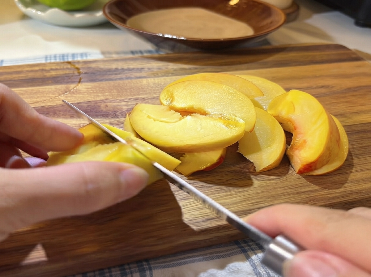 Una mano che usa un piccolo coltello per affettare a spicchi sottili le metà di pesca gialla fresca su un tagliere di legno.