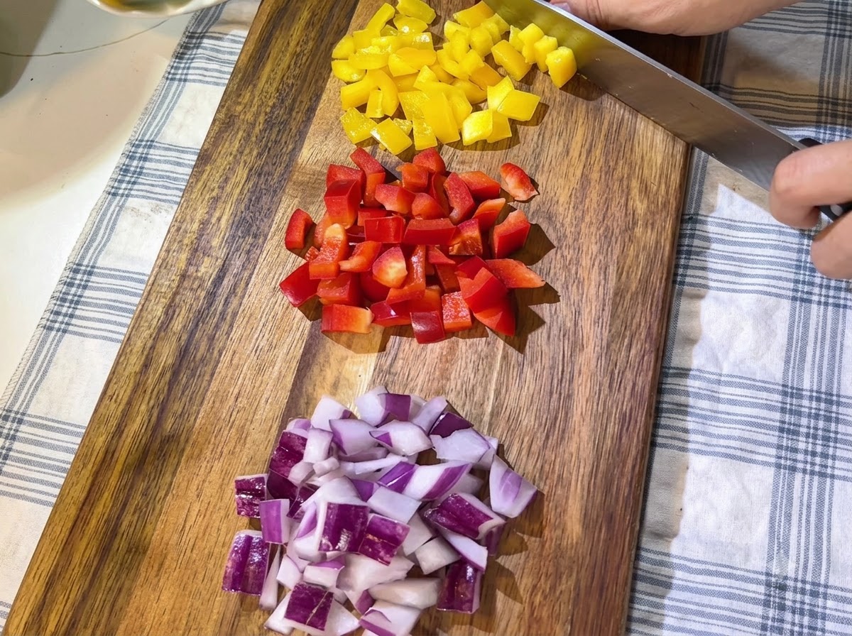 Mucchi di peperoni rossi e gialli freschi tagliati a dadini e cipolle rosse affettate ordinatamente disposti su un tagliere di legno.
