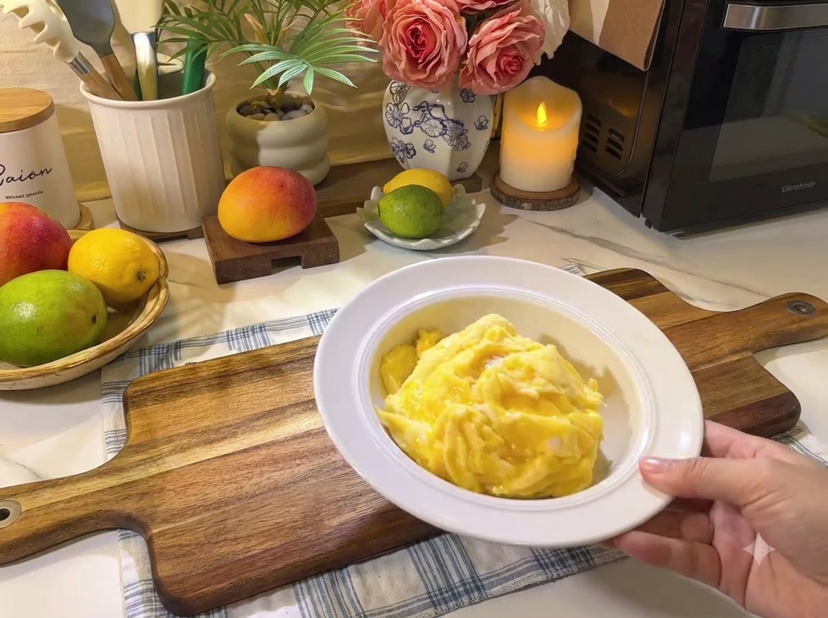 Un piatto di uova strapazzate morbide appoggiato su un asse di legno accanto a frutta fresca e utensili da cucina.