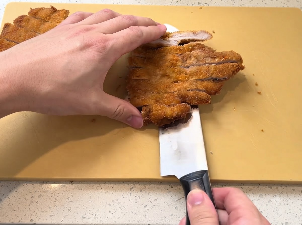 Una cotoletta di maiale fritta dorata tagliata a strisce con un coltello affilato su un tagliere giallo.