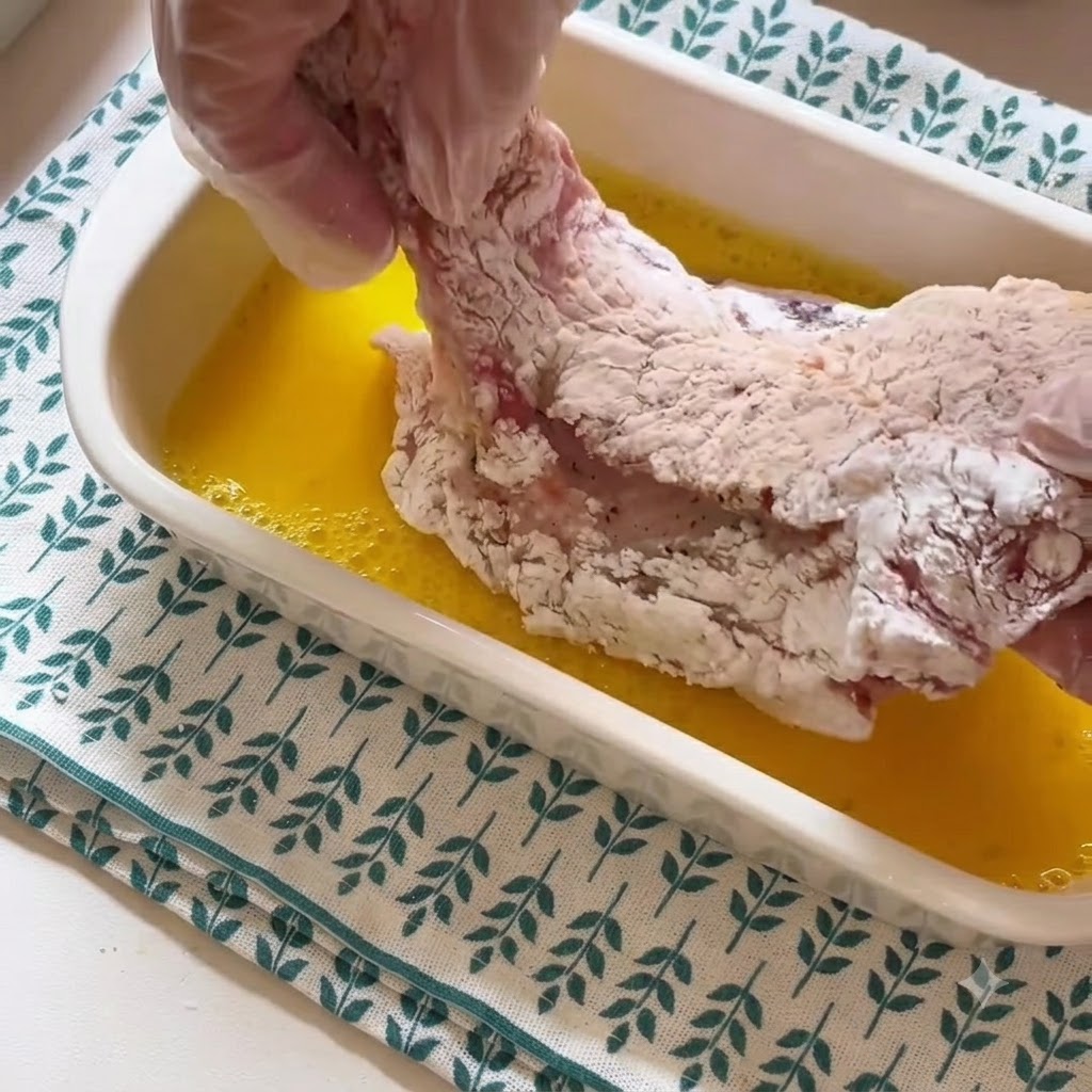 Una mano con il guanto che solleva una cotoletta infarinata da un contenitore di liquido duovo sbattuto giallo.