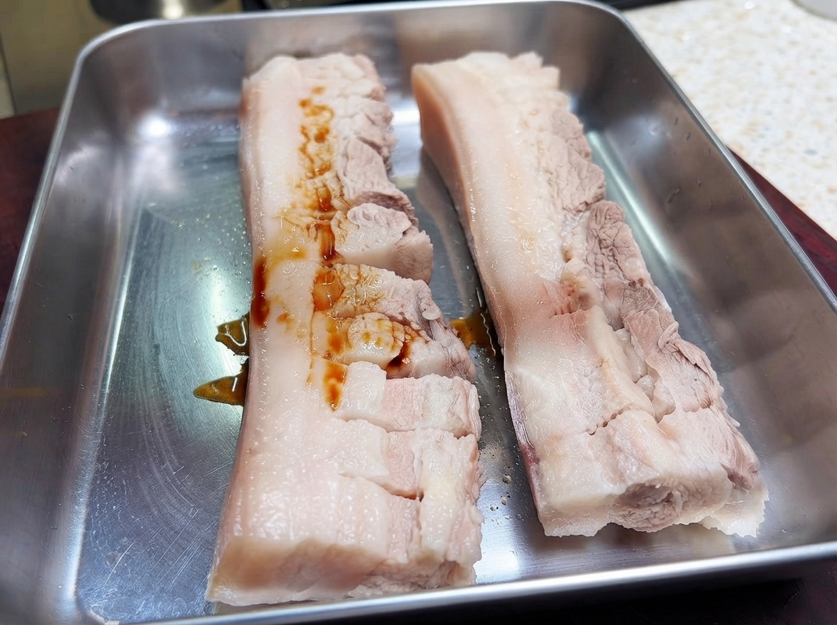 Due pezzi di pancia di maiale incisa su una teglia di metallo con salsa di soia scura versata sulla carne.
