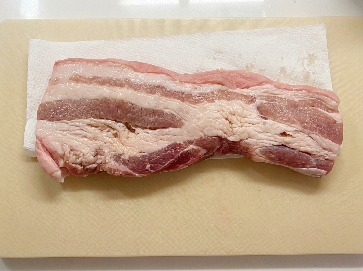 Una striscia di pancetta di maiale sdraiata su un foglio di carta assorbente su un tagliere mentre viene asciugata.
