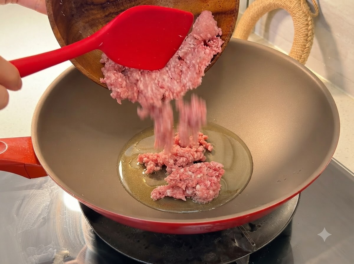 Maiale macinato fresco versato da una ciotola di legno in un wok rosso riscaldato con olio.