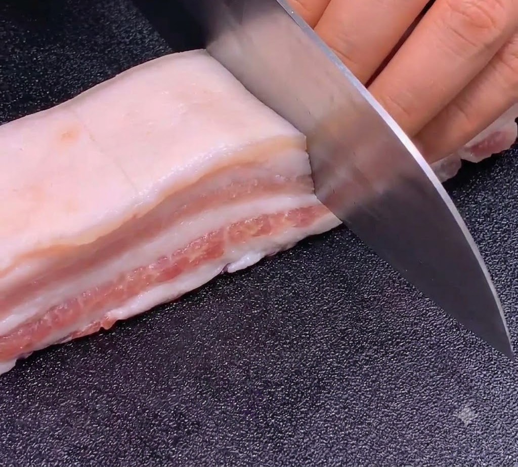 Un coltello affilato che esegue tagli precisi e uniformi su una striscia di pancia di maiale su un tagliere scuro.