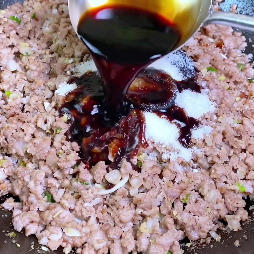Salsa di soia scura versata da un mestolo in un wok contenente carne di maiale macinata e condimenti bianchi.