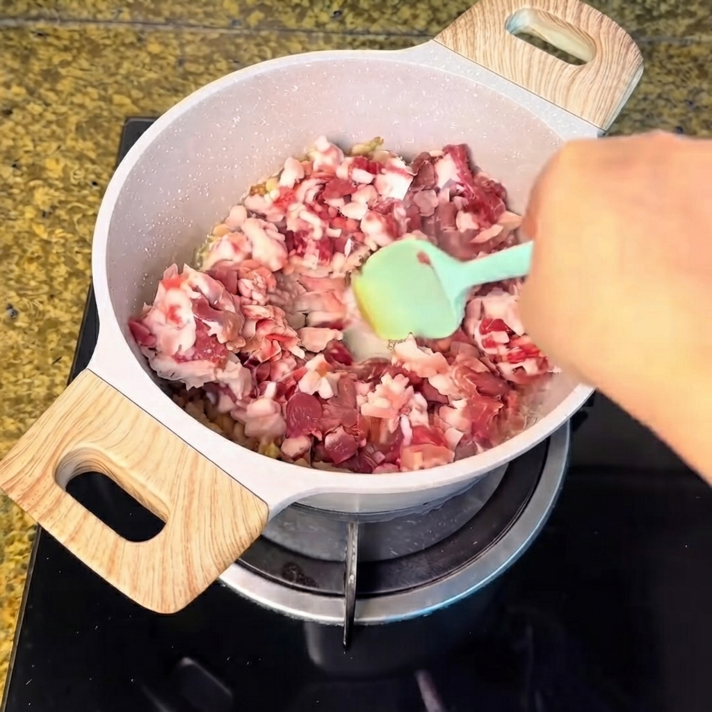 Carne di maiale a cubetti versata nella pentola con il grasso fuso e mescolata con una spatola verde.