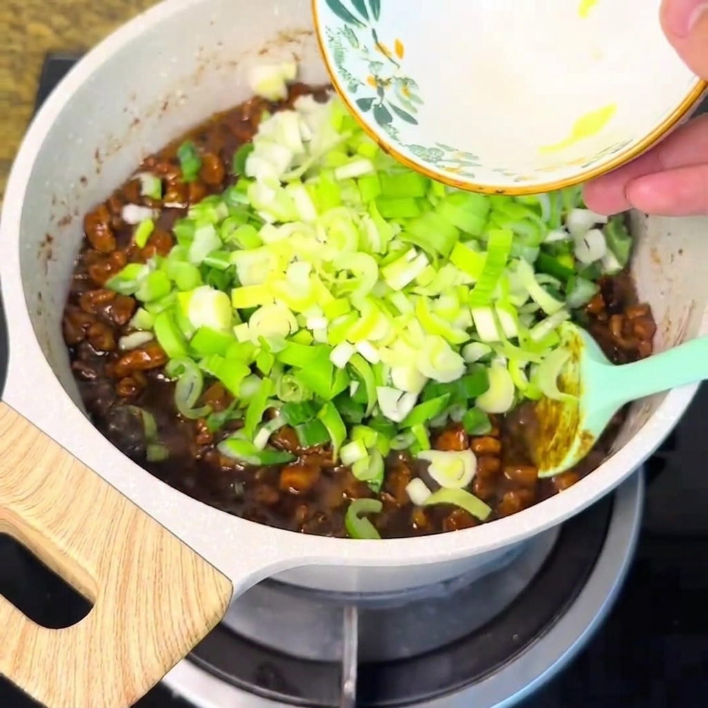 Cipollotti tritati versati da una ciotola nella salsa di carne che sobbolle.