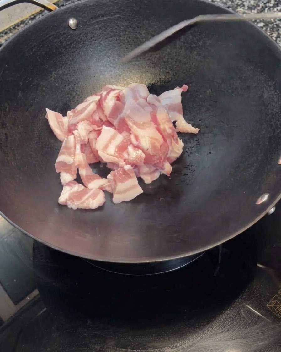 Fette di pancetta di maiale pelata che vengono versate da una ciotola bianca in un wok nero caldo.
