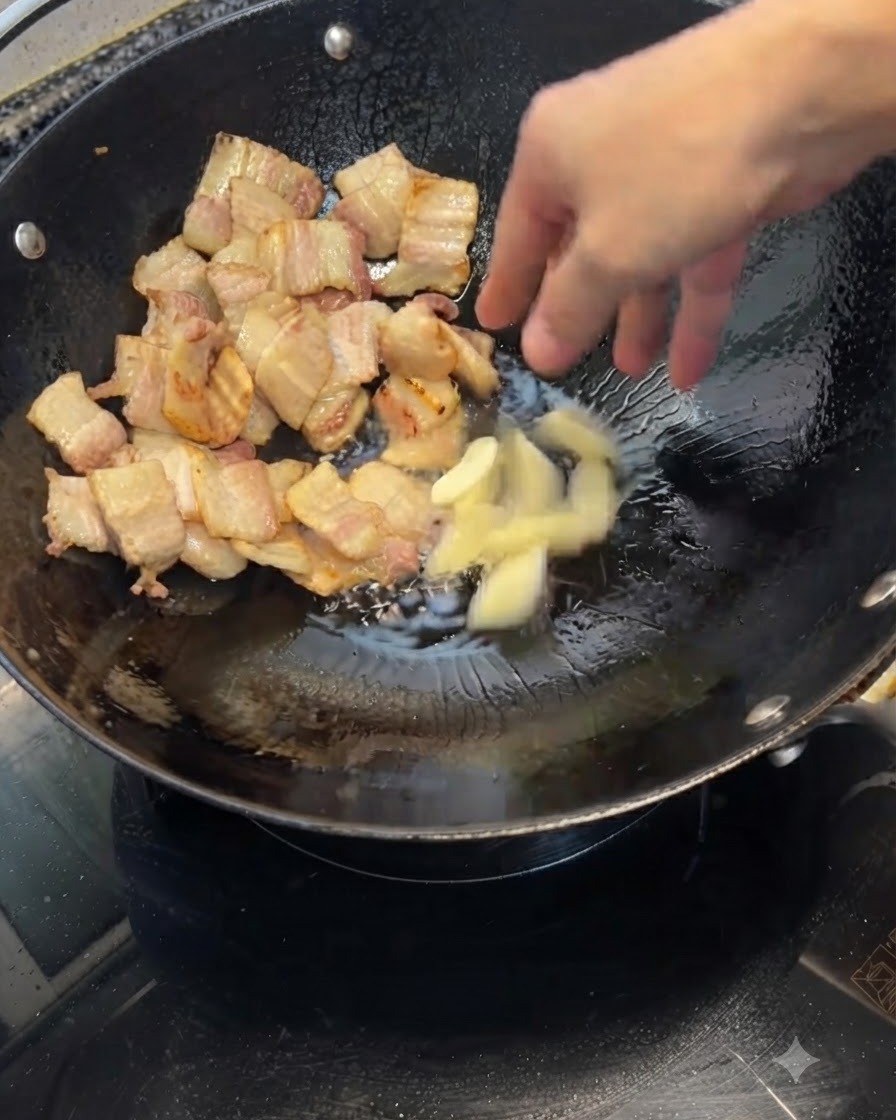 Spicchi daglio schiacciati che vengono gettati in un wok contenente pezzi di pancetta di maiale dorati.