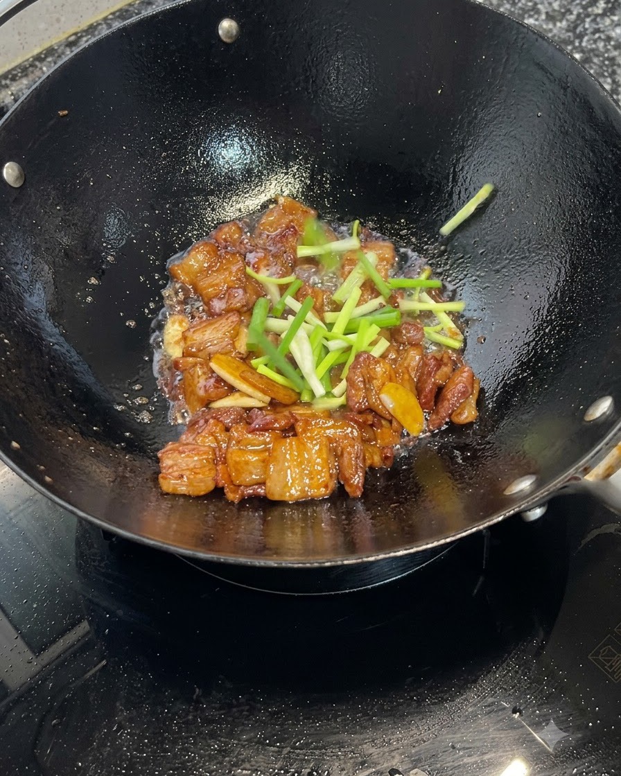 Sezioni di cipollotto fresco sparse sopra pezzi di pancetta di maiale caramellati che cuociono in un wok.