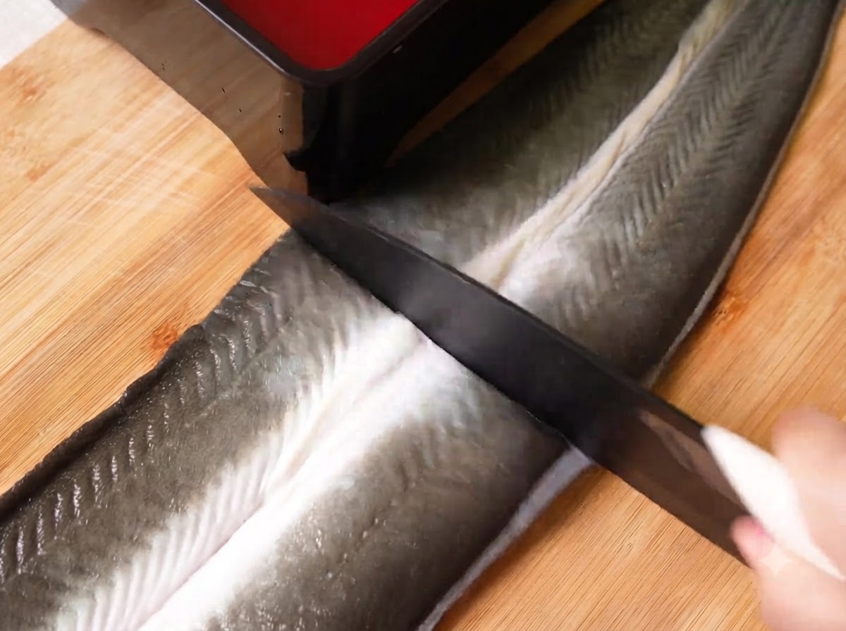 Un coltello affilato che taglia una grande anguilla cruda sventrata in tre sezioni su un tagliere di legno.