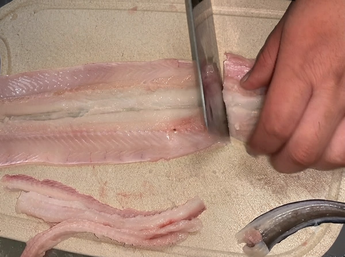 Un coltello che taglia un filetto di anguilla cruda disossata in due sezioni separate su un tagliere.