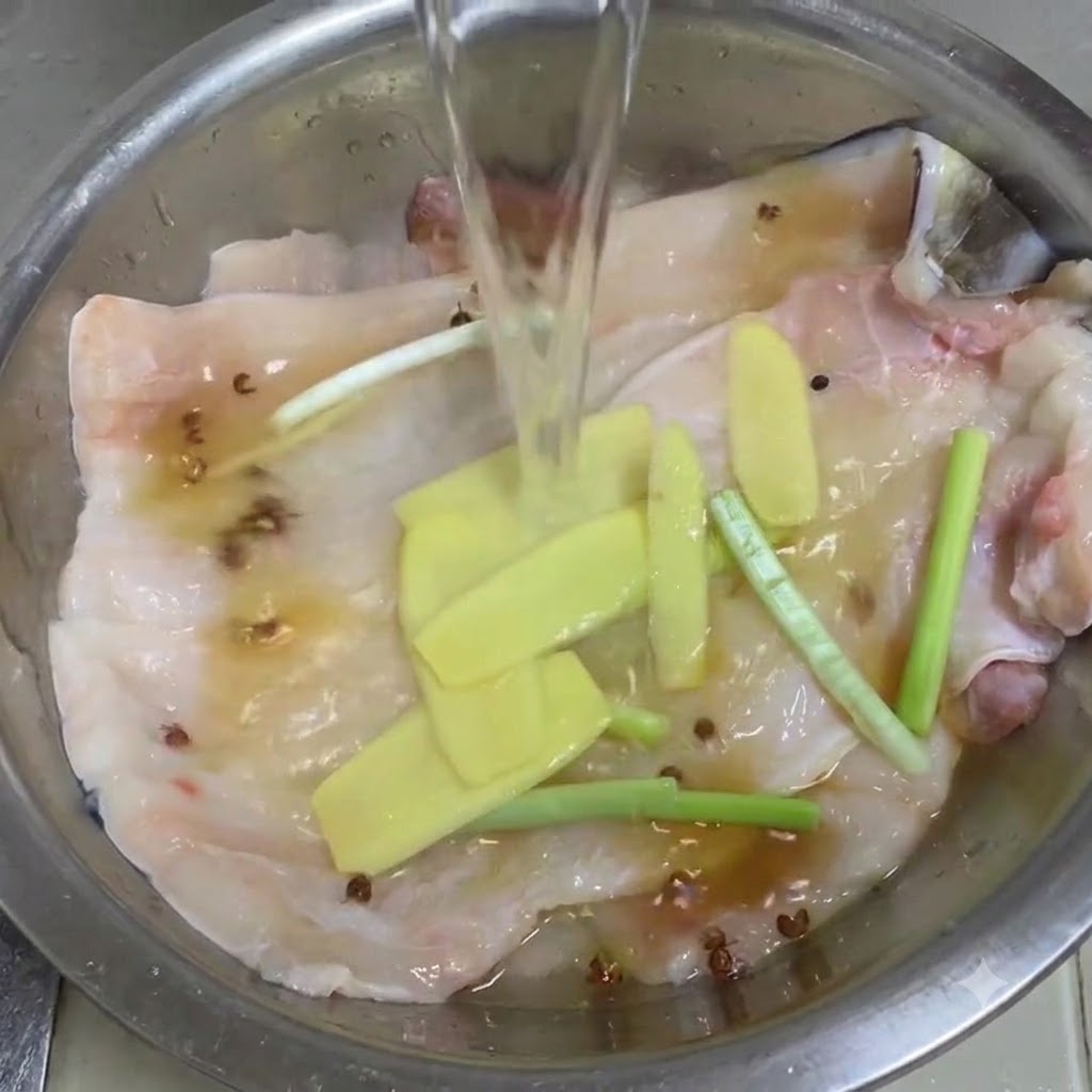 Acqua limpida versata in una ciotola di metallo contenente filetti di anguilla, zenzero, cipollotti e pepe di Sichuan.