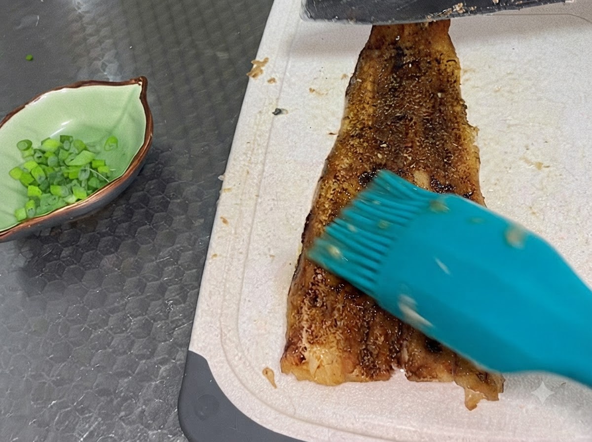 Un filetto di anguilla cotta tagliato su un tagliere bianco accanto a una ciotola verde piena di cipollotti tritati.