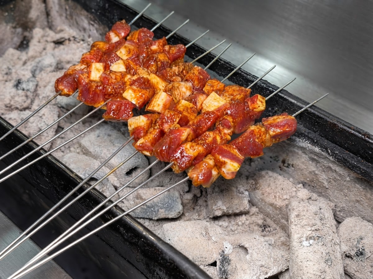 Molteplici spiedini di metallo con carne di maiale condita disposti ordinatamente sopra un barbecue a carbonella ardente.