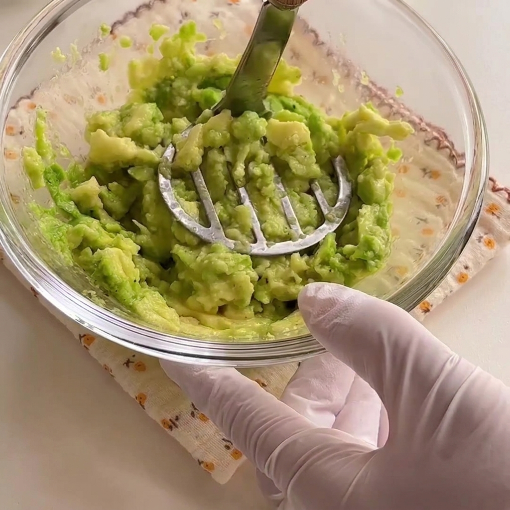 Metà di avocado schiacciate in una pasta verde brillante allinterno di una ciotola di vetro con uno schiacciapatate in metallo.