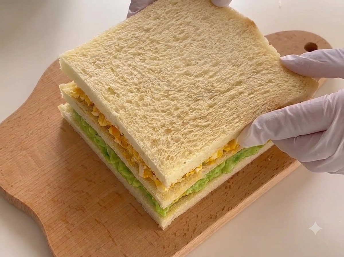 Il panino a tre strati completamente assemblato adagiato su un tagliere, pronto per essere affettato.