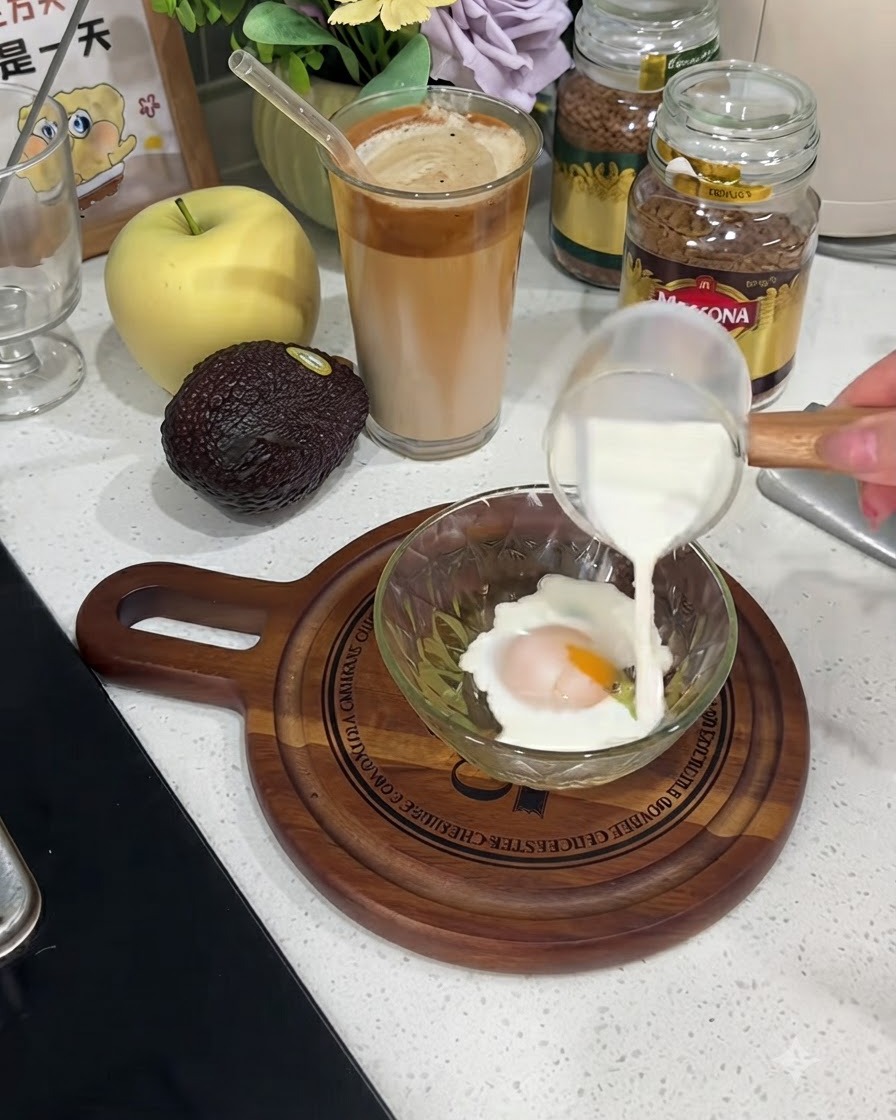 Il latte che viene versato da una piccola lattiera di vetro in una ciotola contenente un uovo rotto.