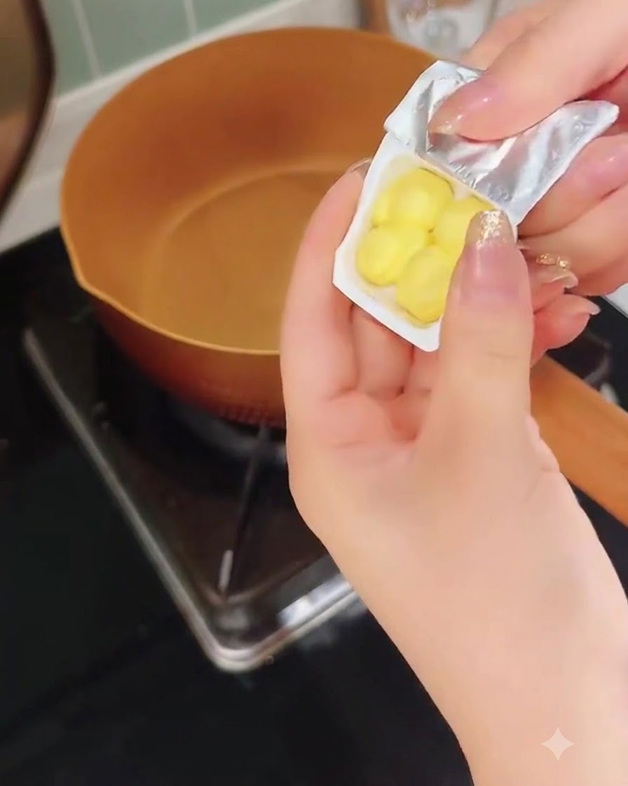 Una mano che sbuccia un piccolo contenitore di plastica con pezzi di burro giallo sopra una padella di rame calda.