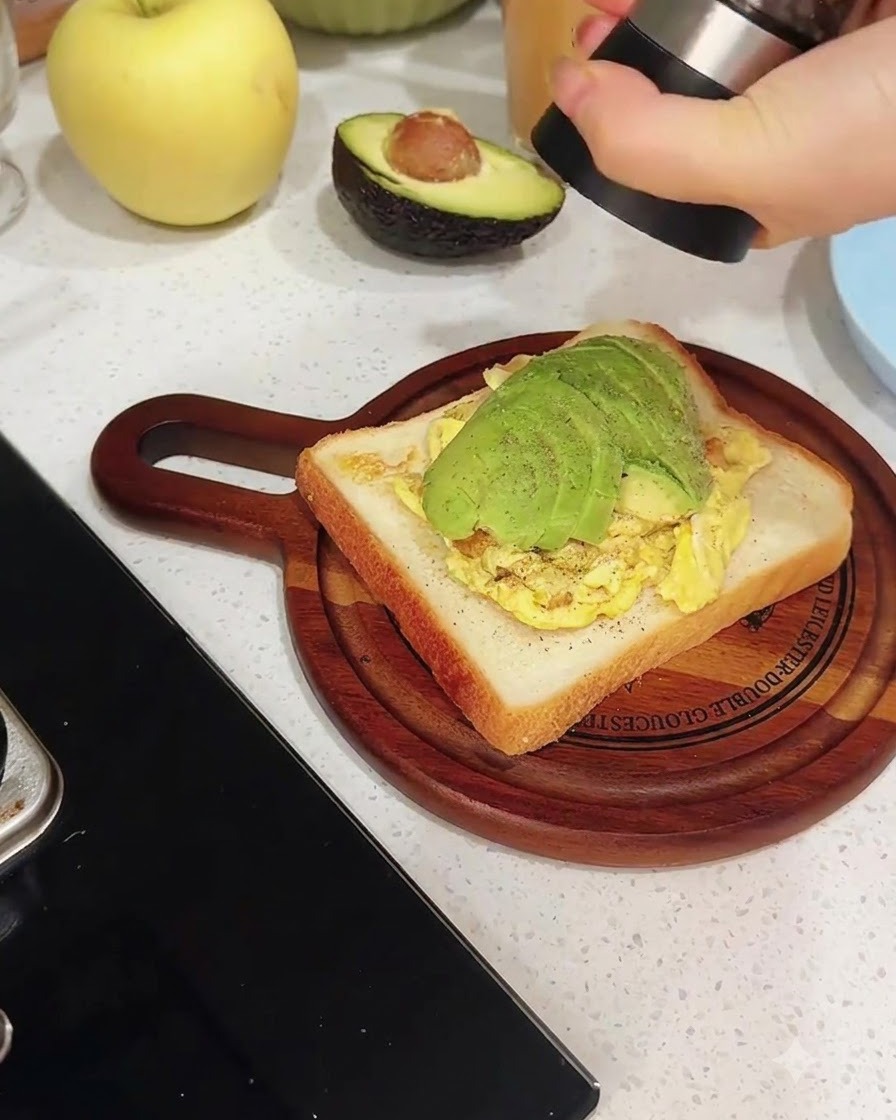 Un macinapepe che macina pepe nero fresco direttamente su una fetta di toast stratificata con uova strapazzate e avocado affettato.