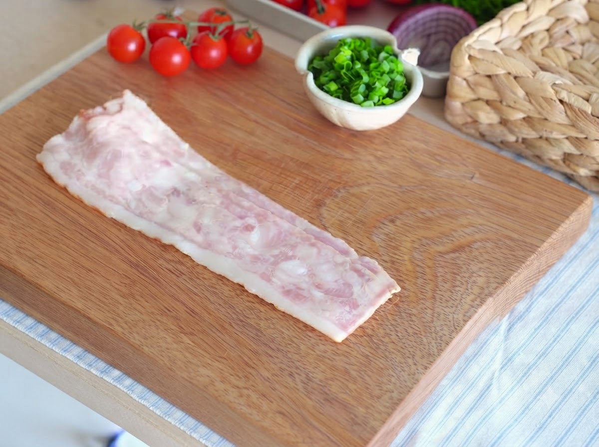 Una singola fetta di pancetta cruda adagiata su un tagliere di legno pronta per essere affettata.