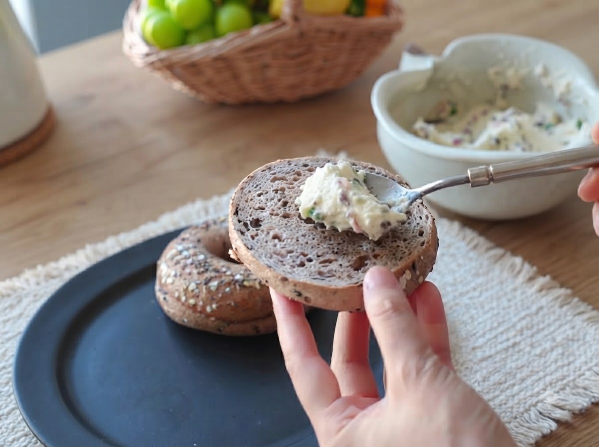 Un cucchiaio di crema di pancetta e cipollotti che viene spalmato sulla superficie di una metà di bagel.