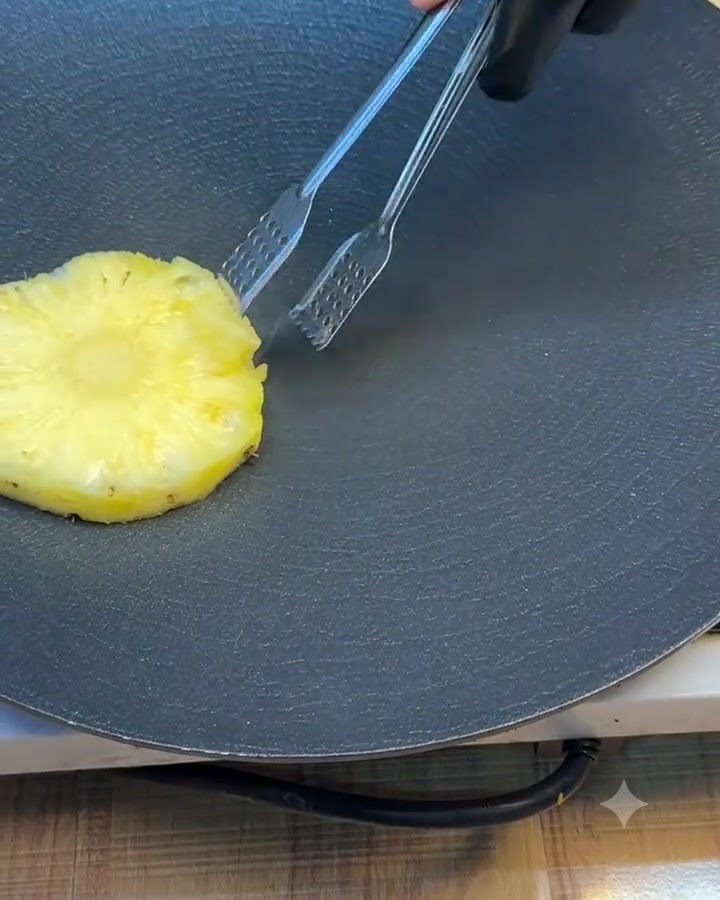 Una singola fetta di ananas girata su una piastra scura con pinze in metallo.