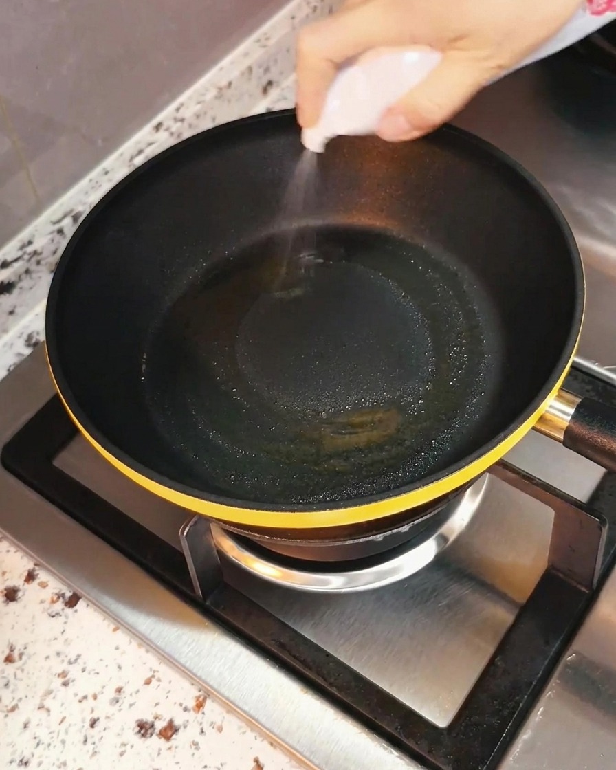 Una mano che spruzza olio da cucina in modo uniforme in una padella nera posizionata su un fornello.