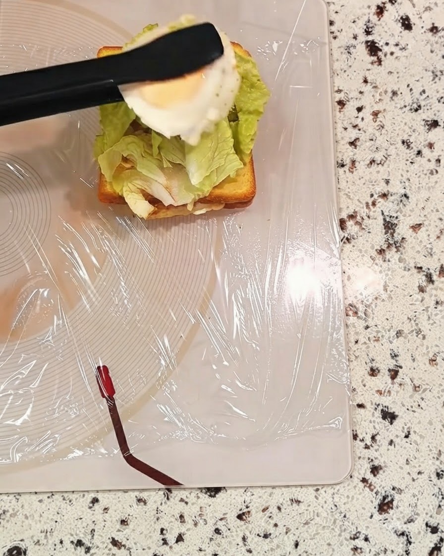 Un paio di pinze nere che tengono un uovo cotto sopra uno strato del panino con linsalata.