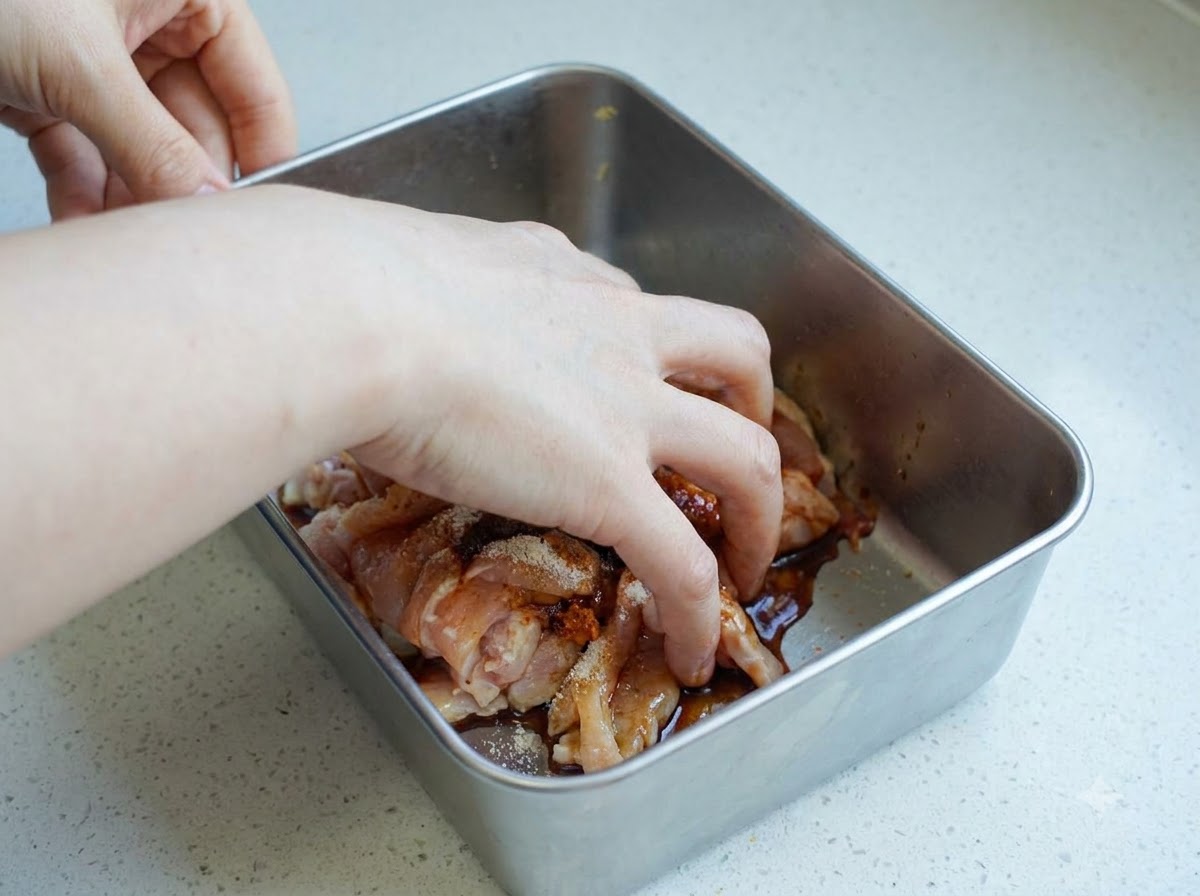 Una mano che mescola fettine di pollo crudo condito allinterno di un contenitore di metallo.