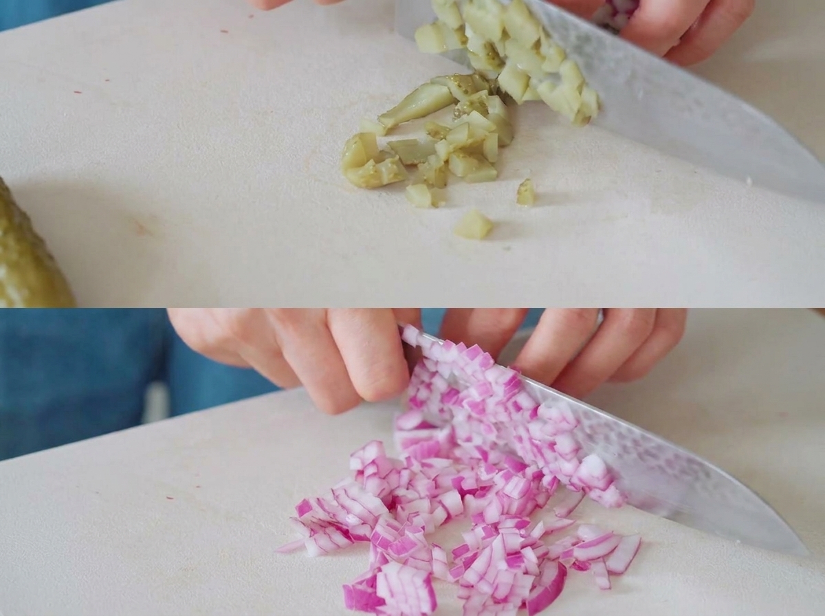 Cetriolini e cipolle rosse tagliati a piccoli pezzi su un tagliere bianco con un coltello da chef.