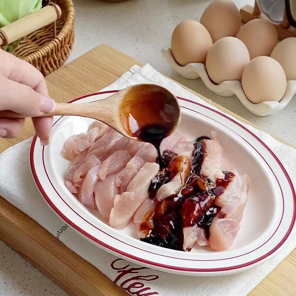 Salse versate da un cucchiaio di legno su strisce di pollo crudo disposte su un piatto bianco.