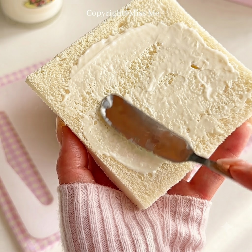Una mano che tiene un coltello per spalmare uniformemente il condimento bianco per insalata su una fetta quadrata di pane bianco.