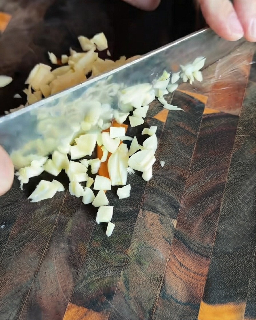 Spicchi daglio freschi tritati finemente e sparsi su un tagliere di legno scuro.