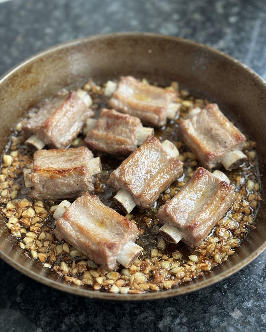 Costine di maiale sbollentate che rosolano in una padella circondate da aglio fritto e olio.