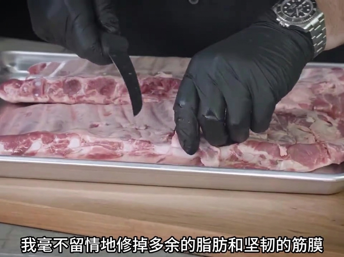 Un paio di mani guantate che usano un coltello per rifilare il grasso dalle costine crude su un vassoio di metallo.
