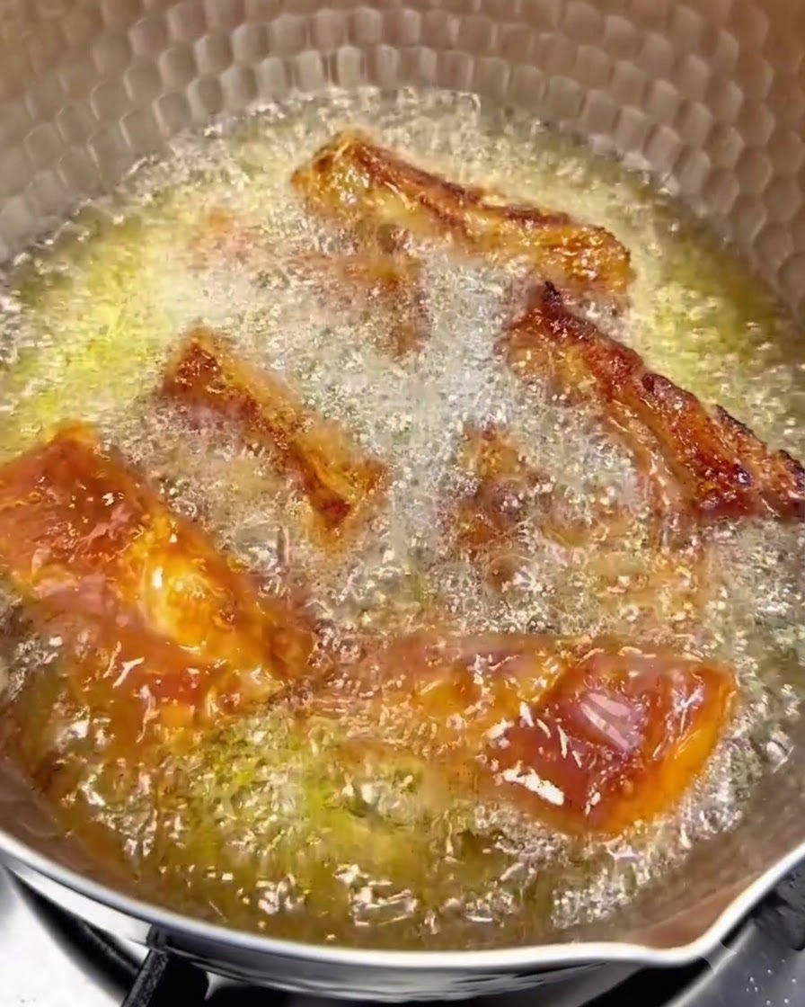 Costine di maiale che friggono nellolio caldo a fuoco basso con superfici dorate visibili.