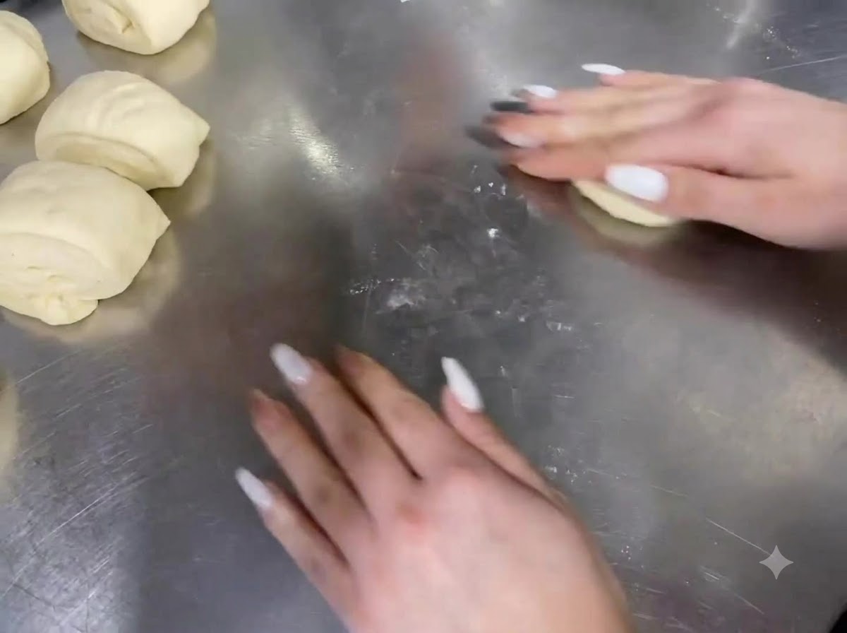 Mani che arrotolano porzioni di impasto su un piano di lavoro in acciaio inossidabile.