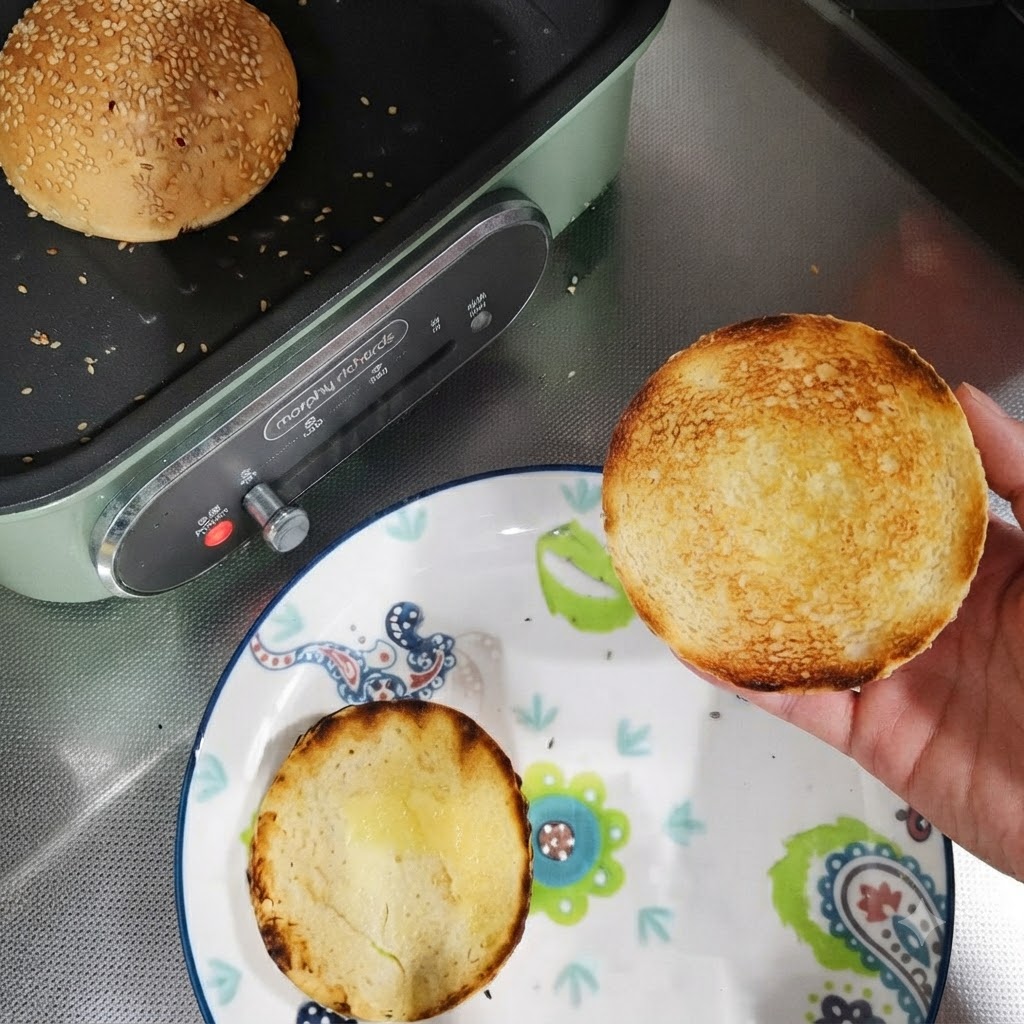 Metà di panini da hamburger tostati su un piatto accanto a una padella con un panino intero in tostatura.