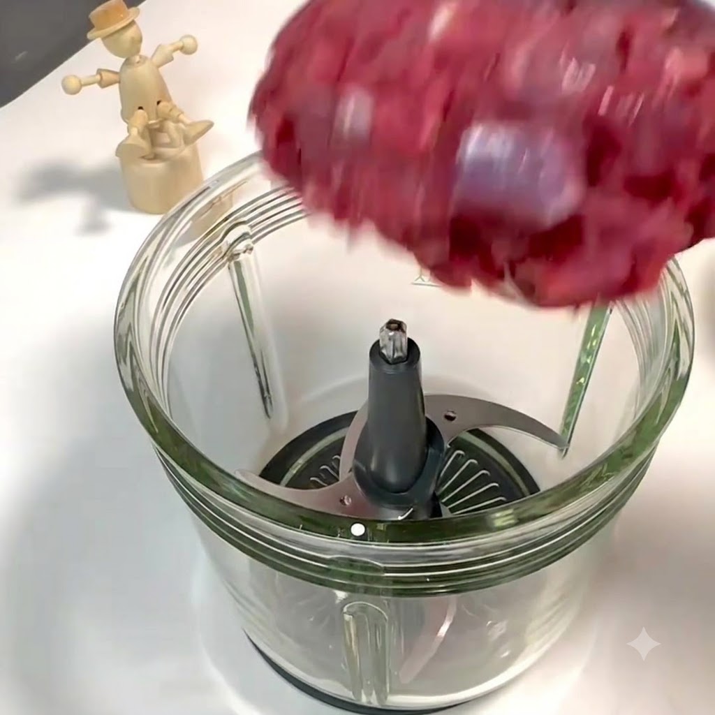 Pezzi di lombo di manzo e pancia di maiale che cadono nel contenitore di vetro aperto di un robot da cucina sopra le lame di metallo.