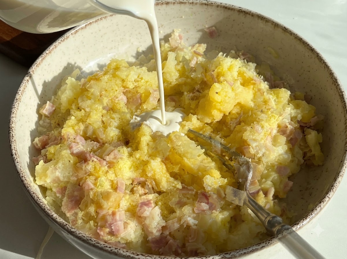 Panna densa versata in una ciotola contenente patate schiacciate e prosciutto a dadini.