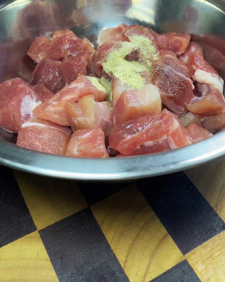 Cubetti di maiale crudo tagliati di fresco dentro una ciotola di acciaio inossidabile cosparsi di condimenti e amido di mais.