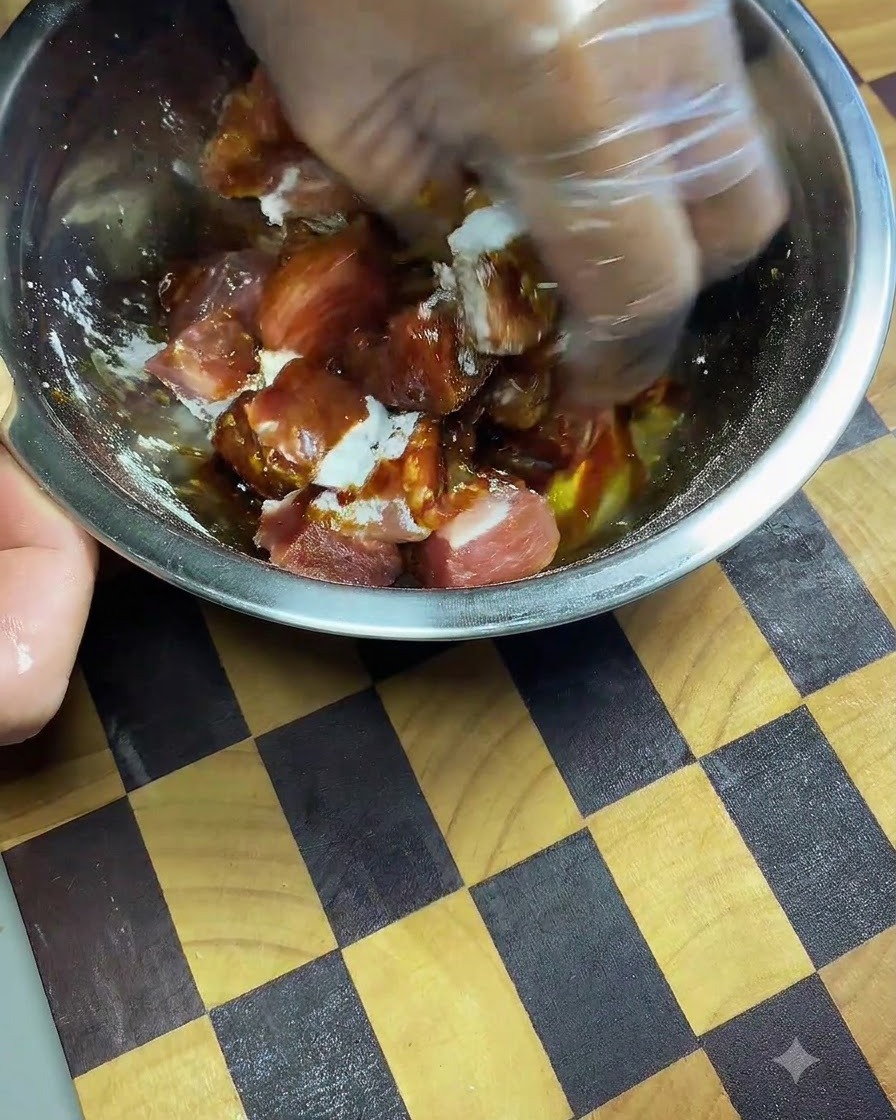 Una mano con il guanto che impasta e massaggia la salsa di soia scura e le spezie nei cubetti di maiale dentro una ciotola di metallo.