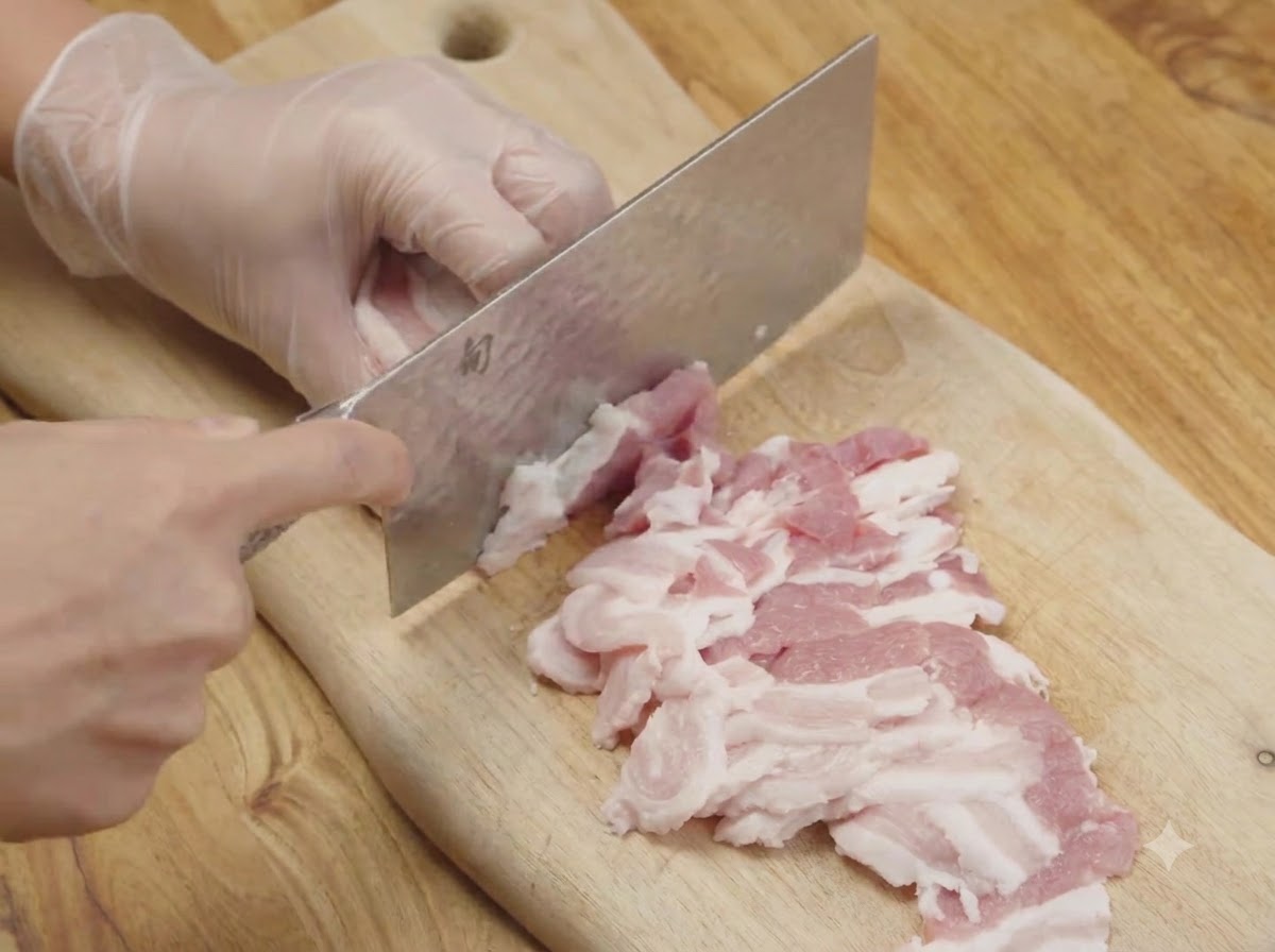 Pancetta di maiale cruda tagliata a strisce sottili su un tagliere di legno usando una mannaia da cucina.