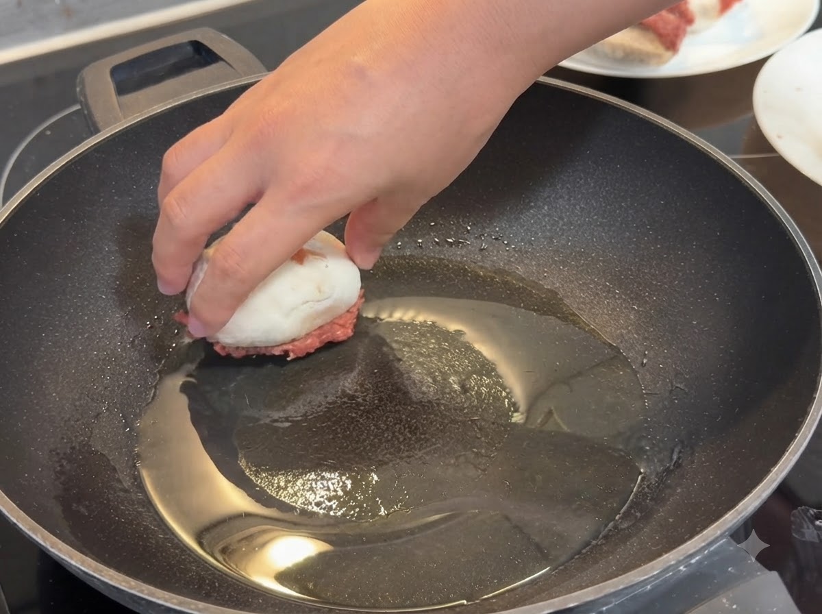 Una mano che posiziona un fungo ripieno con la carne rivolta verso il basso in una padella calda unta dolio.