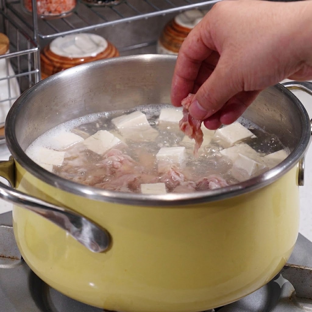 Una mano che inserisce le fette di maiale marinato in una pentola contenente brodo bollente, tofu e frutti di mare.