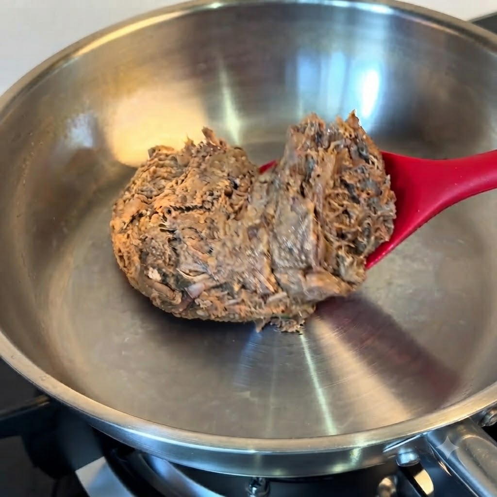 Un blocco di manzo sfilacciato scongelato inserito in una padella in acciaio inossidabile con una spatola rossa.