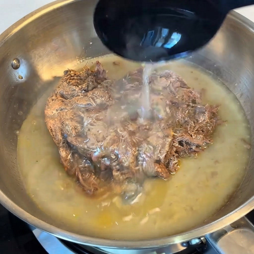 Un mestolo che versa lacqua di cottura amidacea sul manzo sfilacciato in una padella.