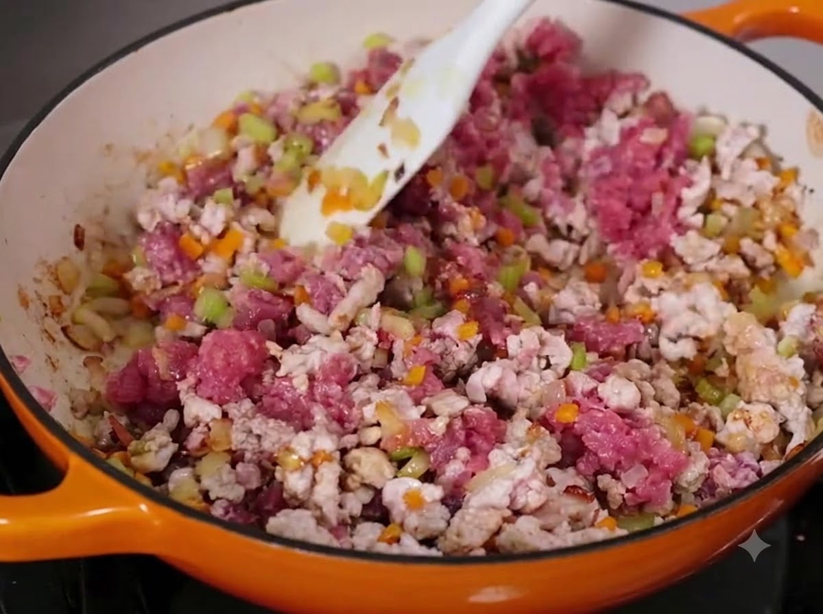 Carne trita di manzo e maiale mescolata con il soffritto in una padella capiente utilizzando una spatola bianca.